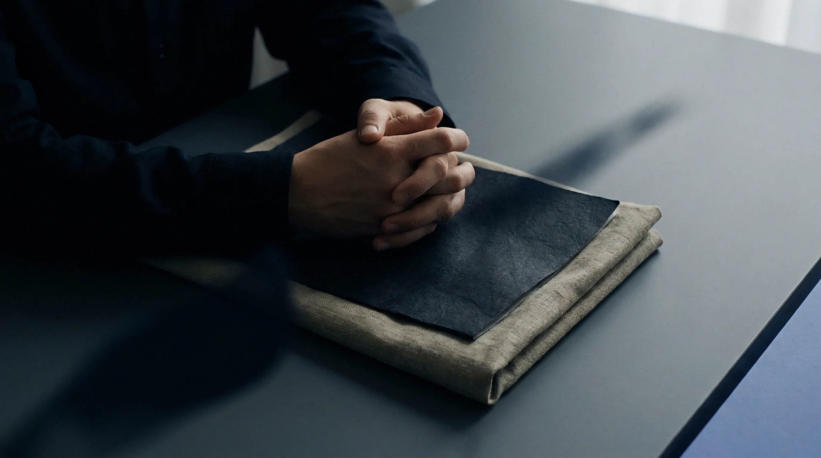 Folded natural flax textile sits beneath a dark fabric layer on a dark tabletop while hands rest near the layered cloth.