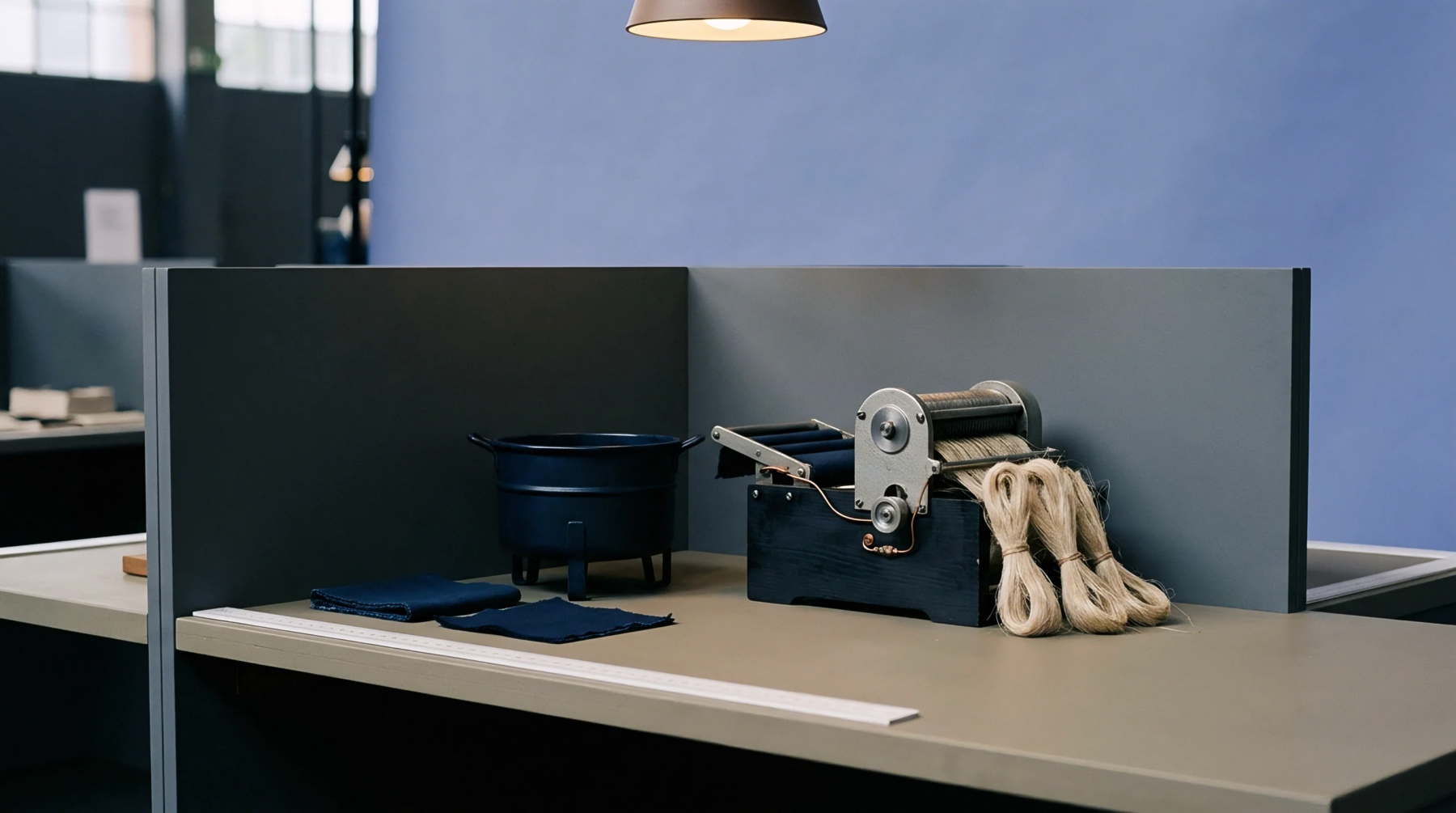 Raw flax fibre hanks rest beside a miniature processing machine and indigo dyed fabric samples on a workshop table.