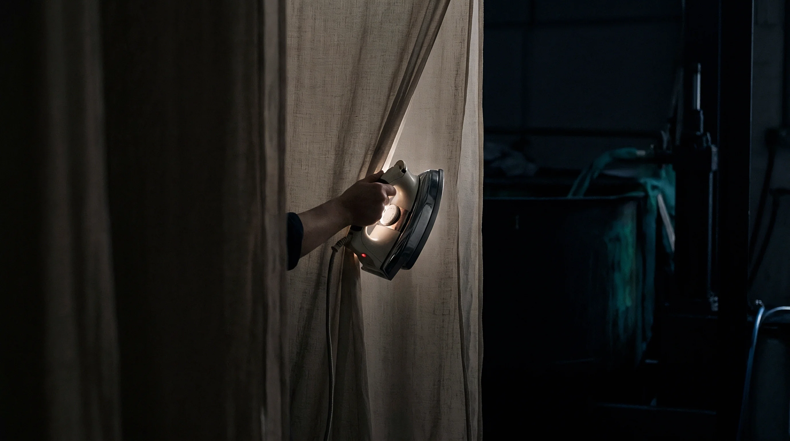 A bare hand guides an electric iron along a hanging panel of woven flax fabric inside a dark workshop.