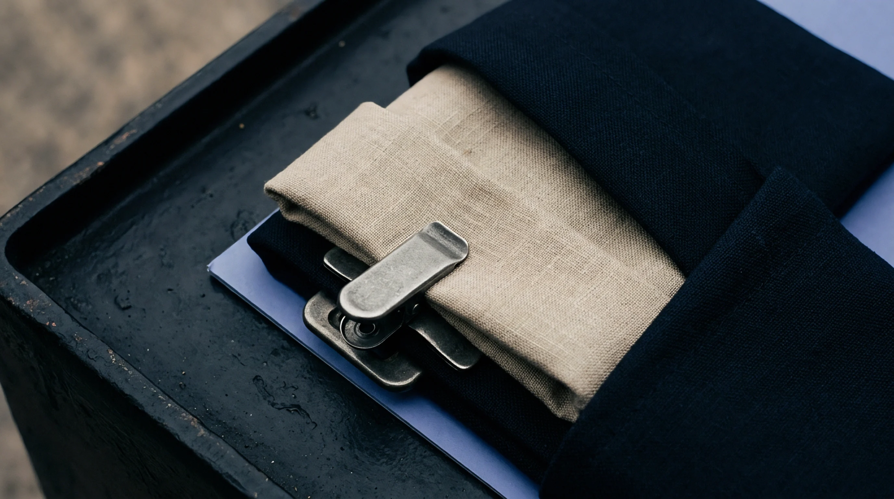 The image shows several neatly folded pieces of woven cloth, likely linen, in natural beige and dark blue shades, secured with metal clips on a dark, textured surface.